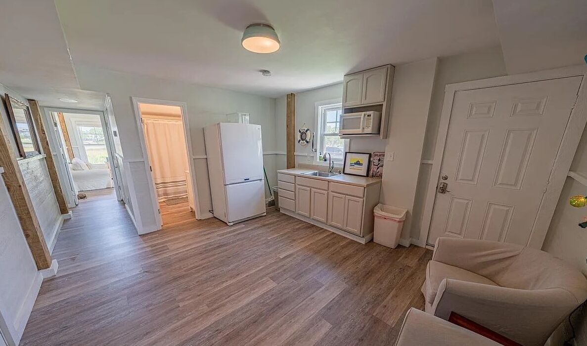 Una cocina compacta con armarios blancos, un refrigerador y un microondas. El piso es de madera y hay una puerta que da a otra habitación.