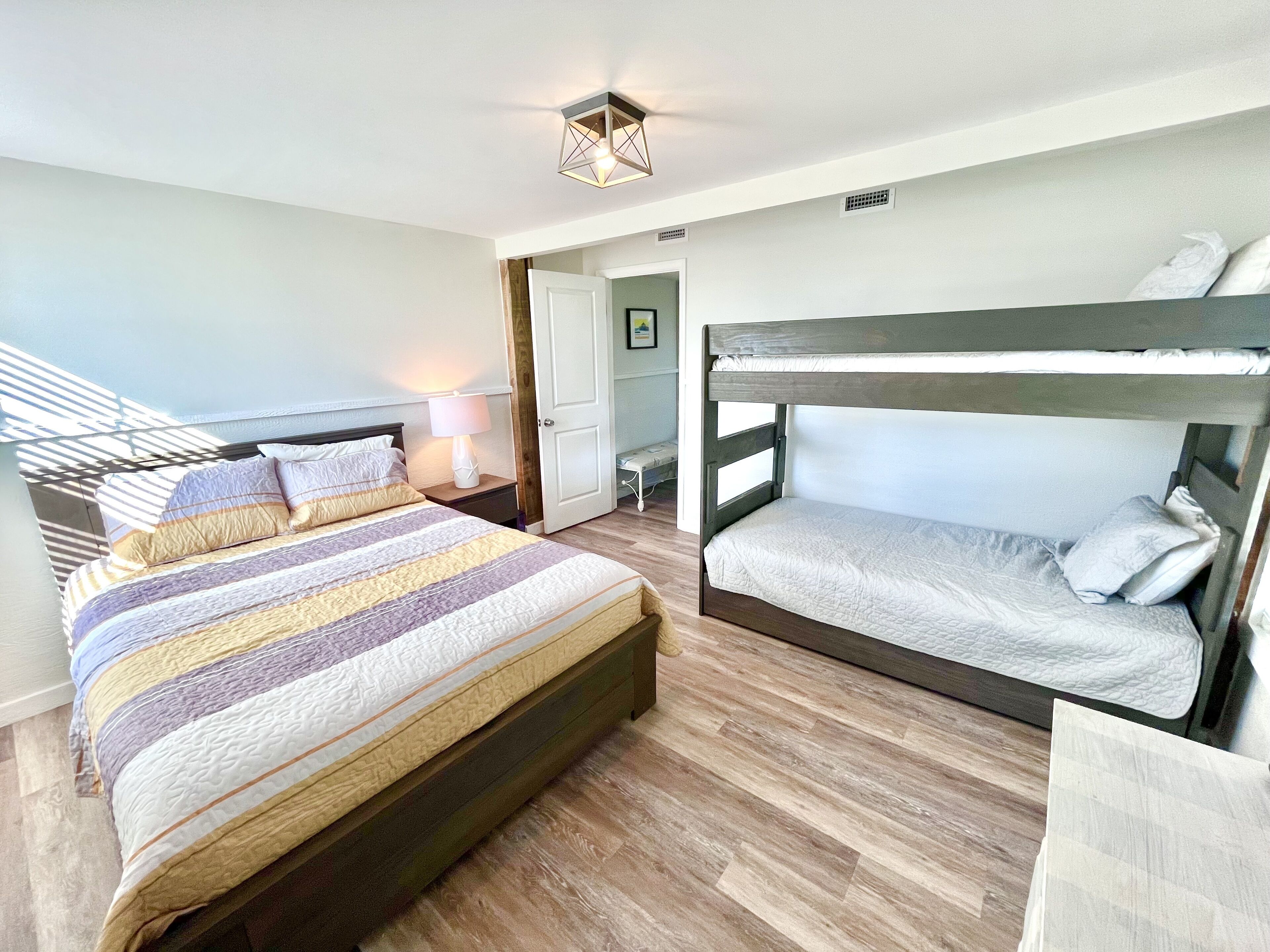 Una habitación con una cama grande, una cama individual superpuesta, una mesita de noche con lámpara y una puerta que da a otra habitación.