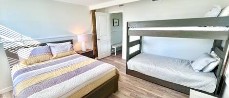 Una habitación con una cama grande, una cama individual superpuesta, una mesita de noche con lámpara y una puerta que da a otra habitación.