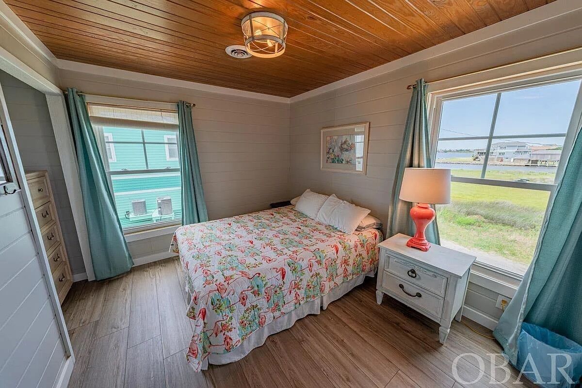 A bedroom with a bed, a nightstand, a lamp, a window with curtains, and a painting on the wall.