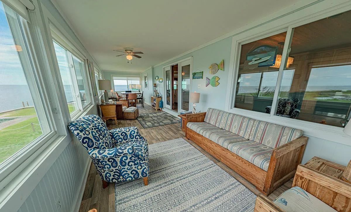A spacious living room with a large window overlooking the ocean, a wooden sofa, patterned armchair, and a ceiling fan.