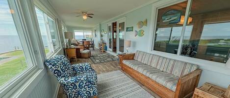 A spacious living room with a large window overlooking the ocean, a wooden sofa, patterned armchair, and a ceiling fan.