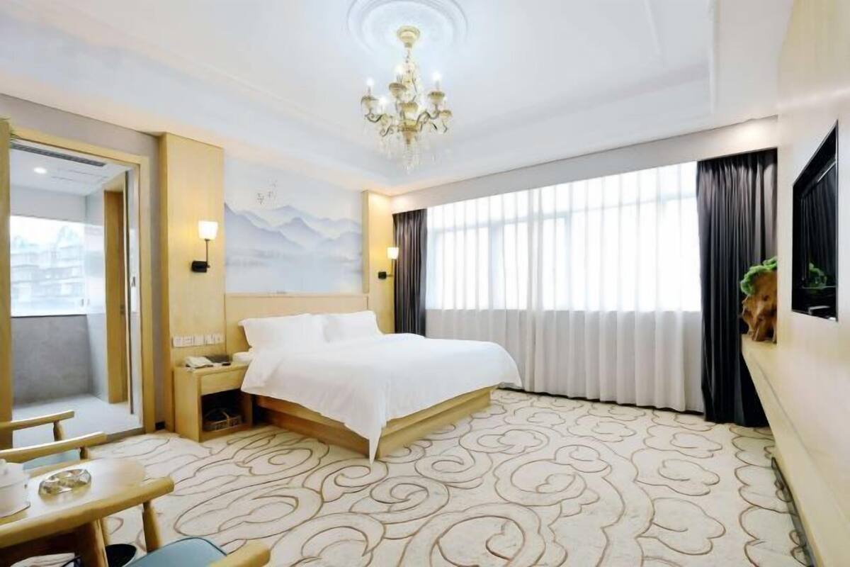 Quarto de hotel com cama, mesa de cabeceira, luminária e vista para a cidade através de grandes janelas.