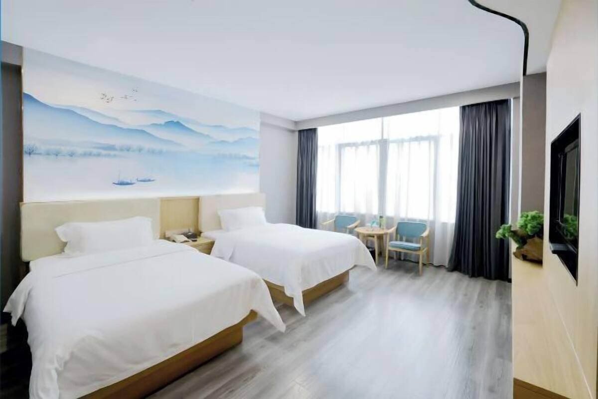 Quarto de hotel com duas camas, uma escrivaninha, uma cadeira, um quadro na parede e uma janela grande com cortinas.