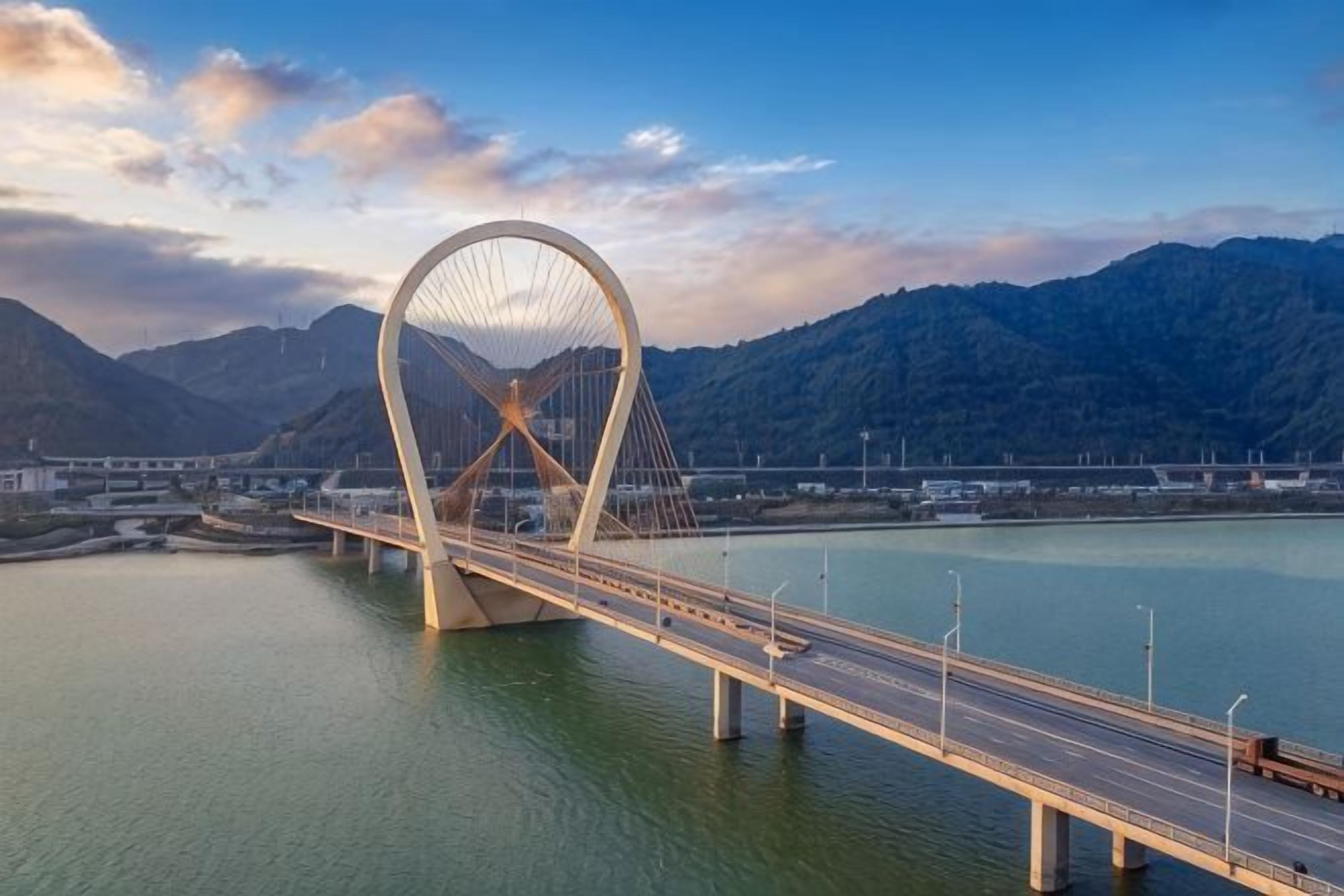 A large bridge with a distinctive arch design spanning a body of water, with mountains in the background.