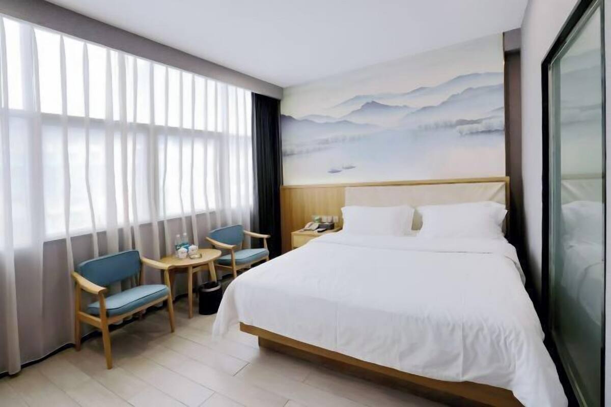 Quarto de hotel com cama, secretária, duas cadeiras, janela com cortinas e um quadro na parede.