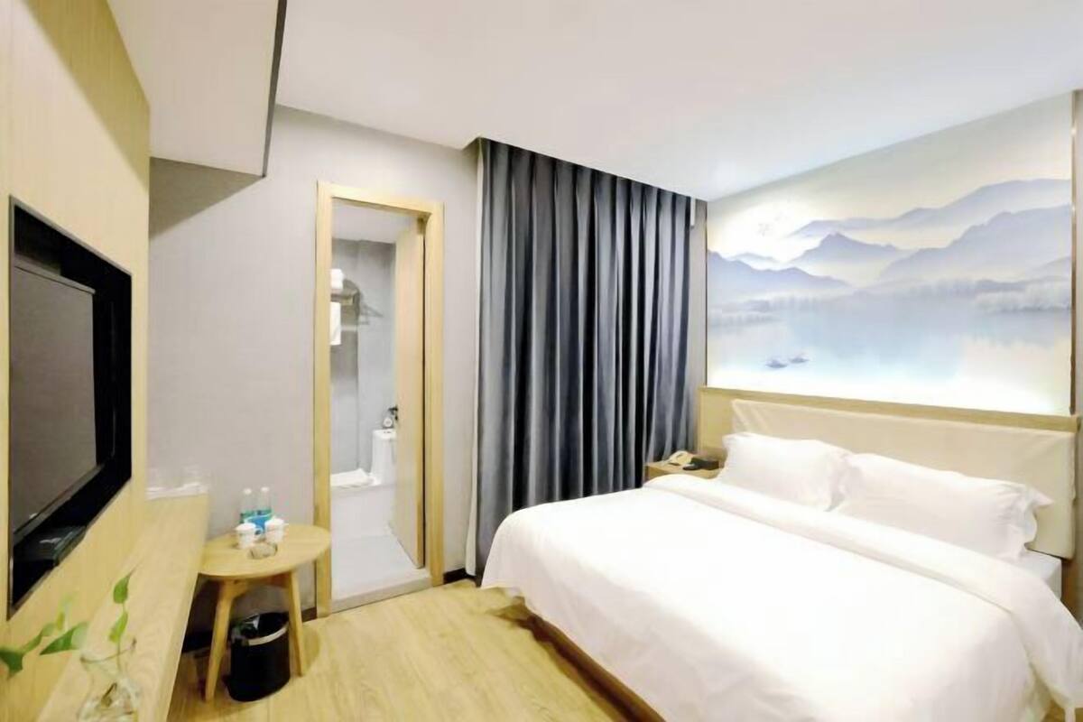 Quarto de hotel com uma cama, televisão, uma pequena mesa com produtos de higiene, espelho e uma pintura de montanhas.