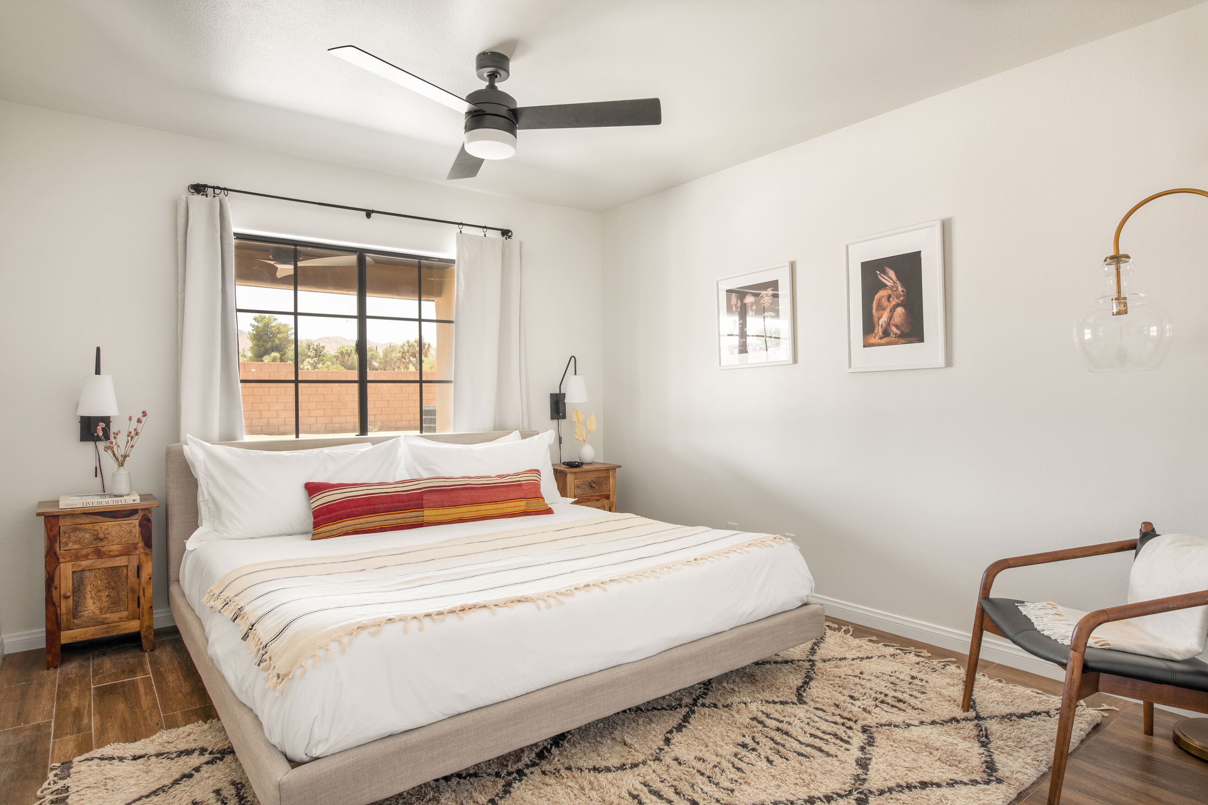 A bedroom with a bed, a ceiling fan, a window with curtains, a nightstand with a lamp, and a chair.