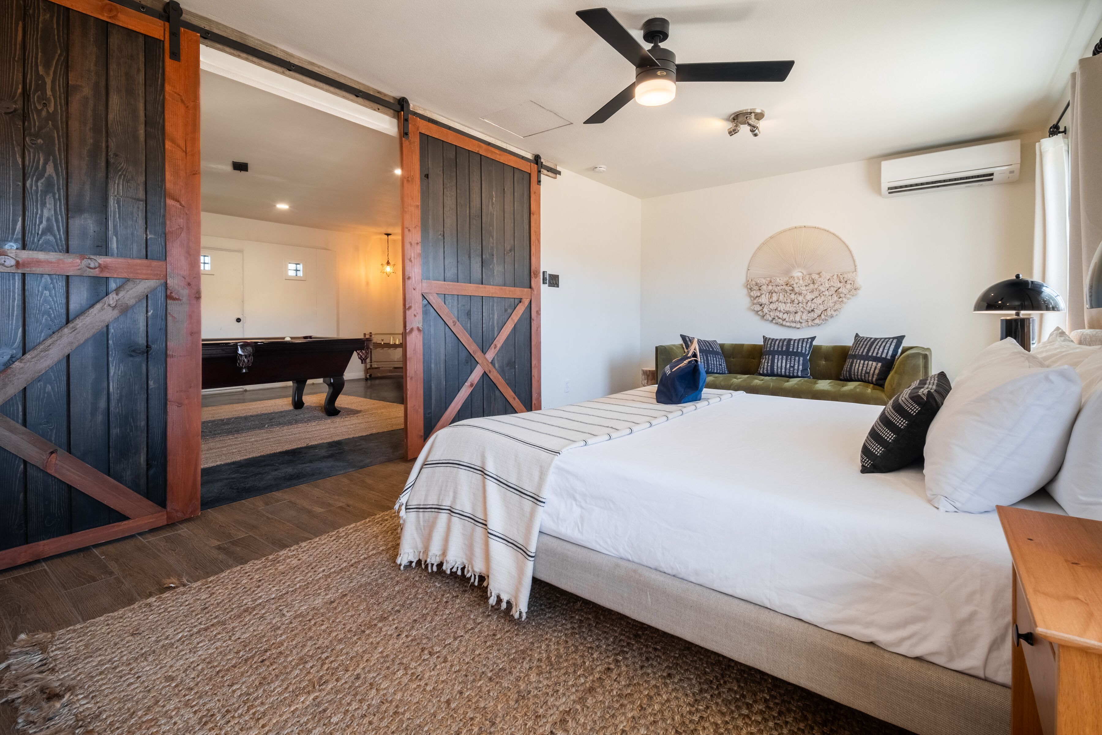 A bedroom with a large bed, a green sofa, a wooden desk, a ceiling fan, and a rustic barn door.
