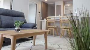 A modern living room with a grey sofa, a wooden coffee table, and a small potted plant. There is a dining area with wooden chairs and a white table. The room has a white carpet and a white wall. - buransiera naha akebono puremisuto 1009 (Naha)