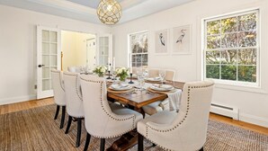 Comedor con una mesa de madera dispuesta para seis personas, rodeada por seis sillas tapizadas con detalles de botones. Se observa un ventanal con vistas a árboles y una lámpara colgante sobre la mesa.