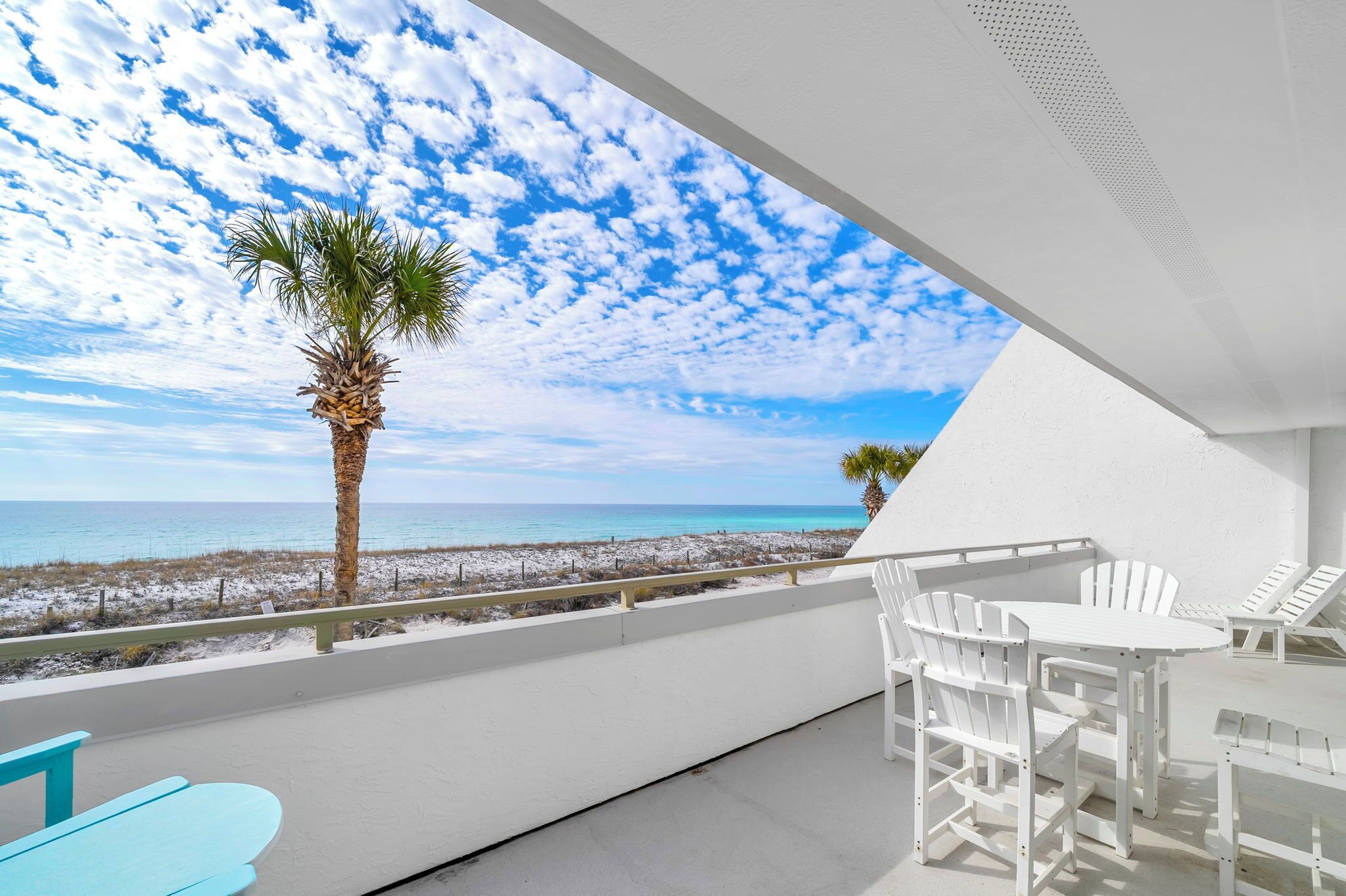 A balcony with a view of the beach and ocean, white furniture, and a palm tree.