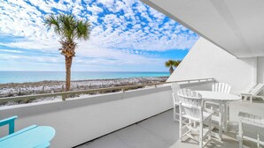 A balcony with a view of the beach and ocean, white furniture, and a palm tree.