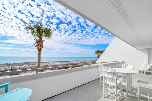 A balcony with a view of the beach and ocean, white furniture, and a palm tree.