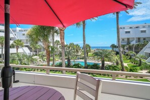 Un balcon agrémenté d’un parasol rouge, donnant sur un espace piscine bordé de palmiers et offrant une vue sur l’océan.