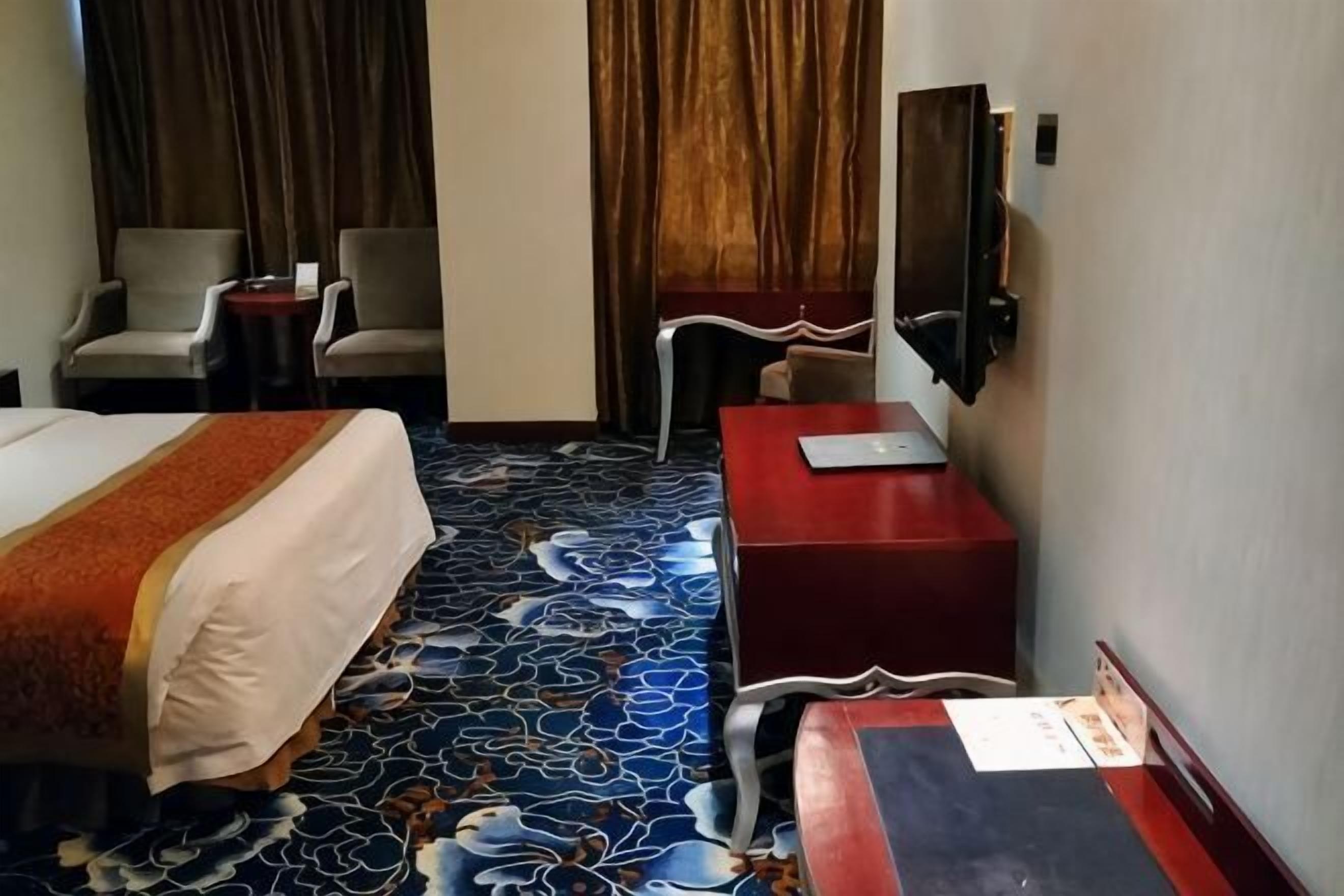 A hotel room with a bed, a desk, a chair, a TV, and a patterned carpet.