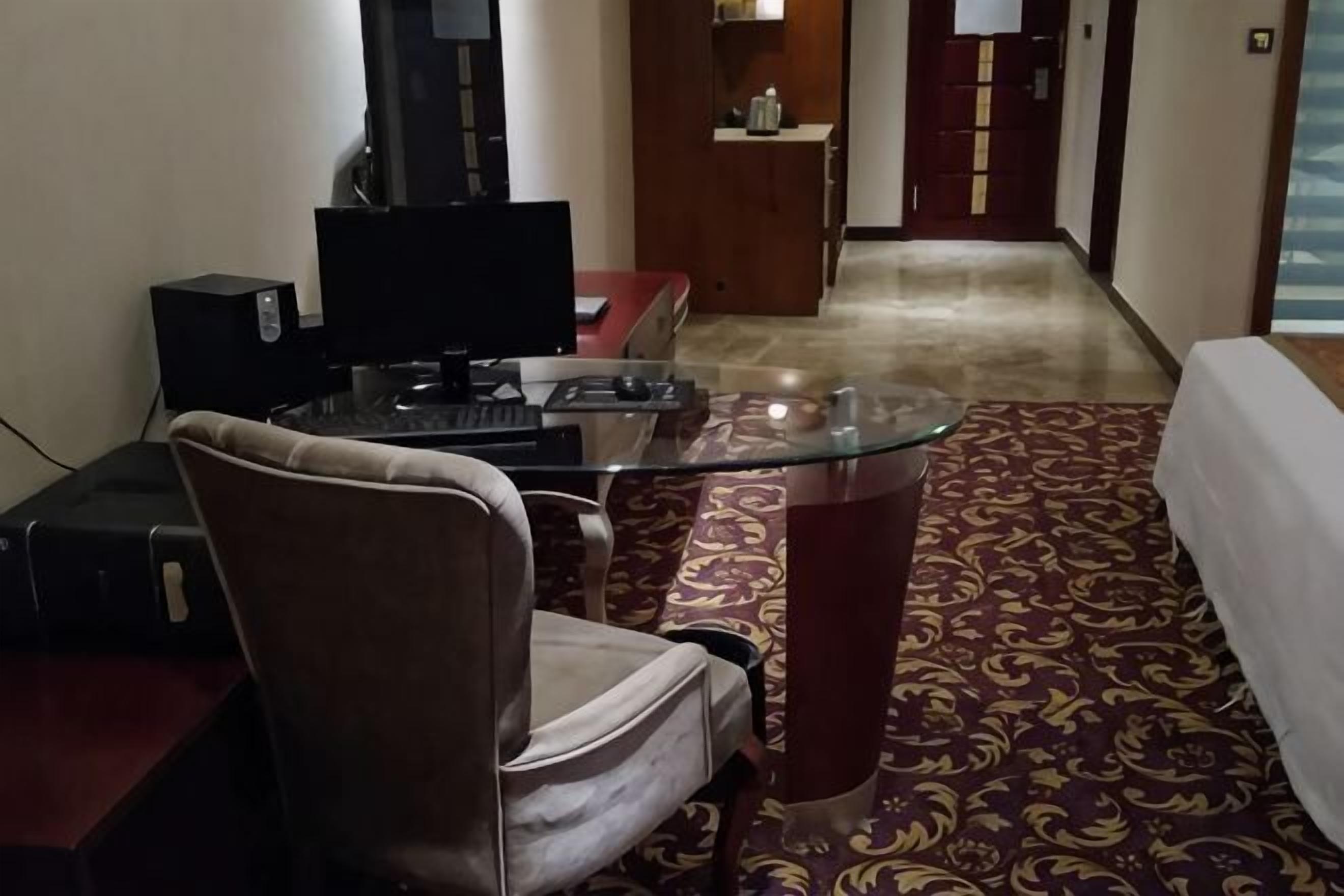 A hotel room with a glass table, a chair, a desk with a computer, a television, and a patterned carpet.