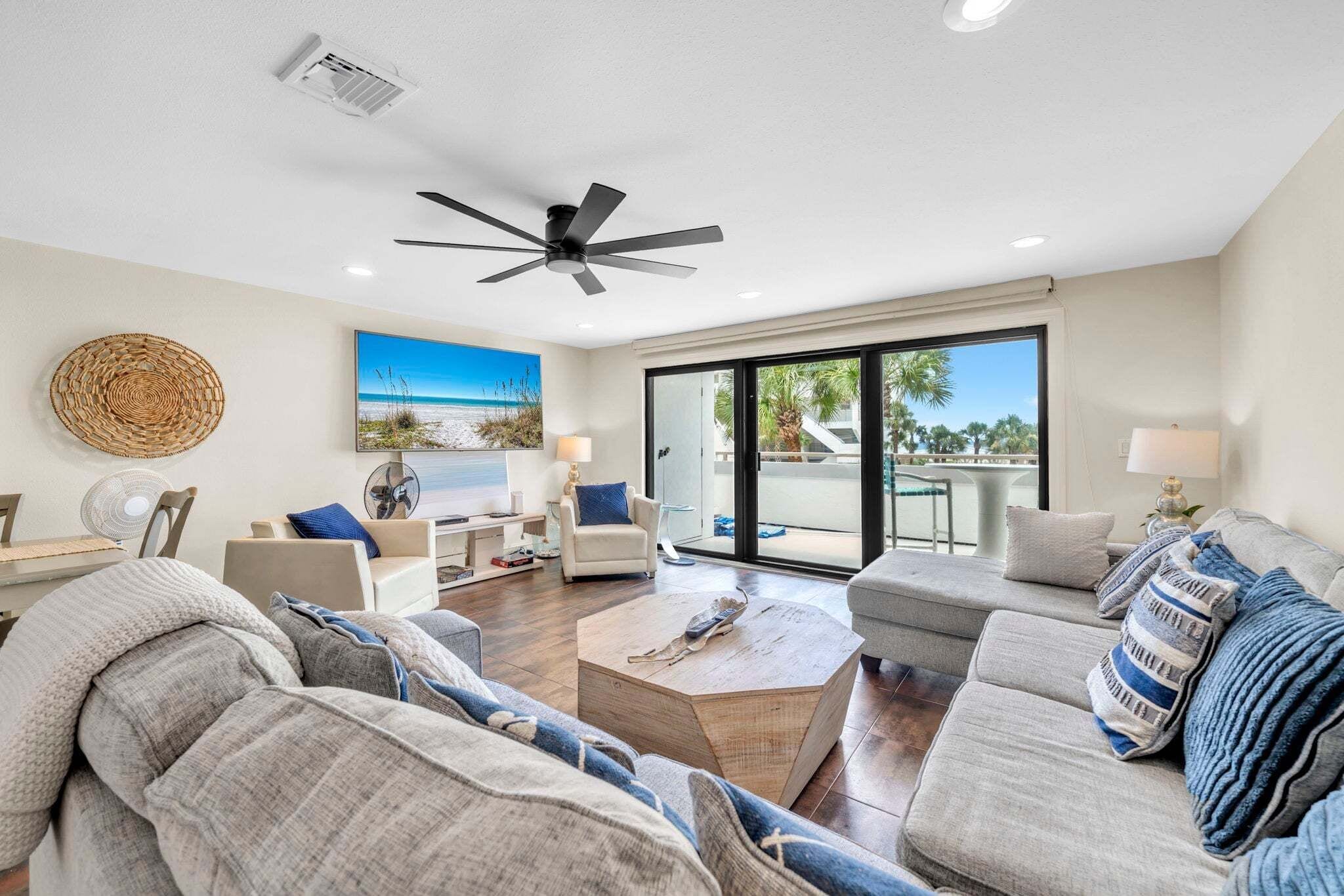 A modern living room with a ceiling fan, a large window with a view of palm trees, and a cozy seating area with a coffee table.