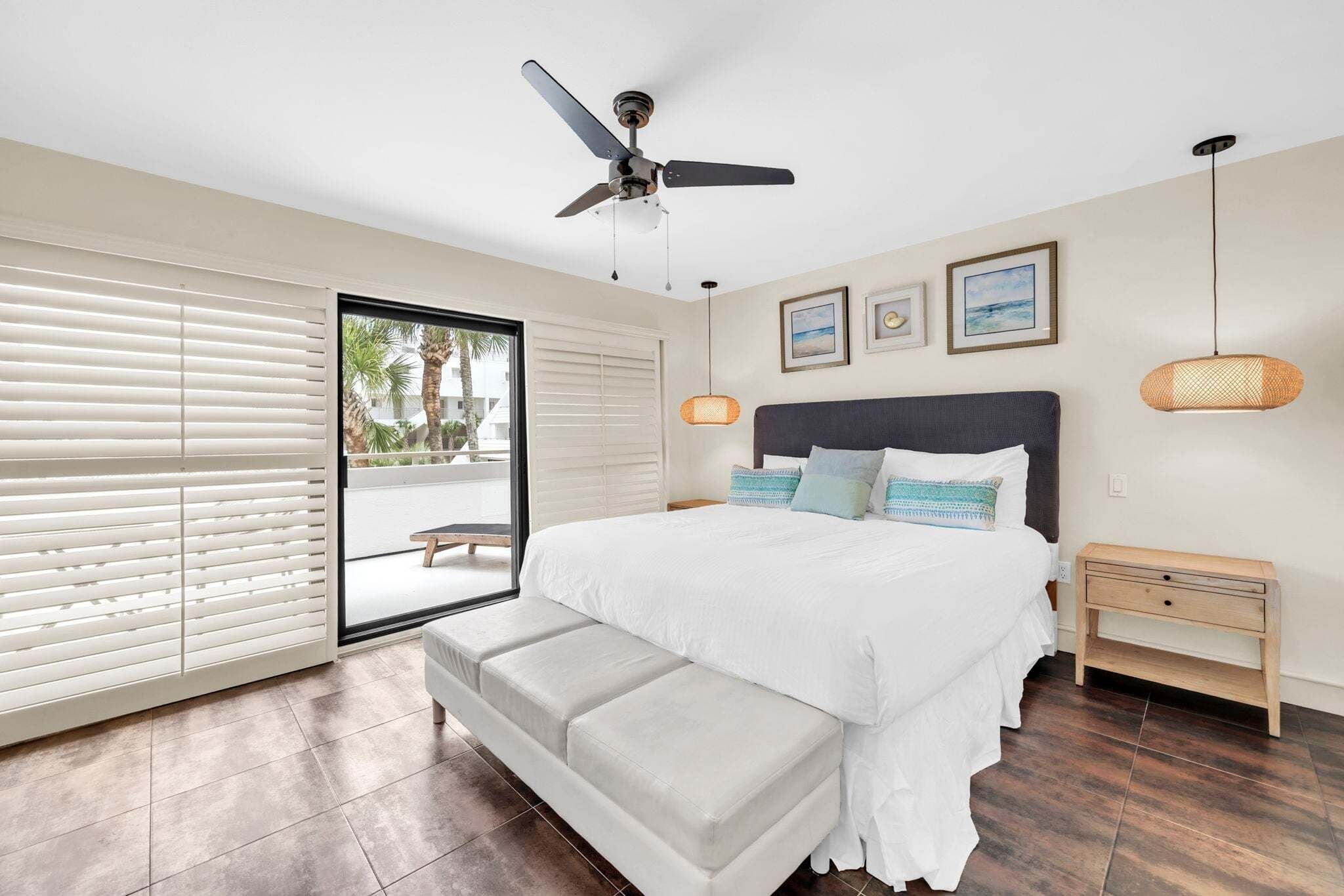 Une chambre à coucher avec un lit, un ventilateur de plafond, un banc, une table de chevet et une vue sur l’extérieur, visible à travers une porte vitrée.