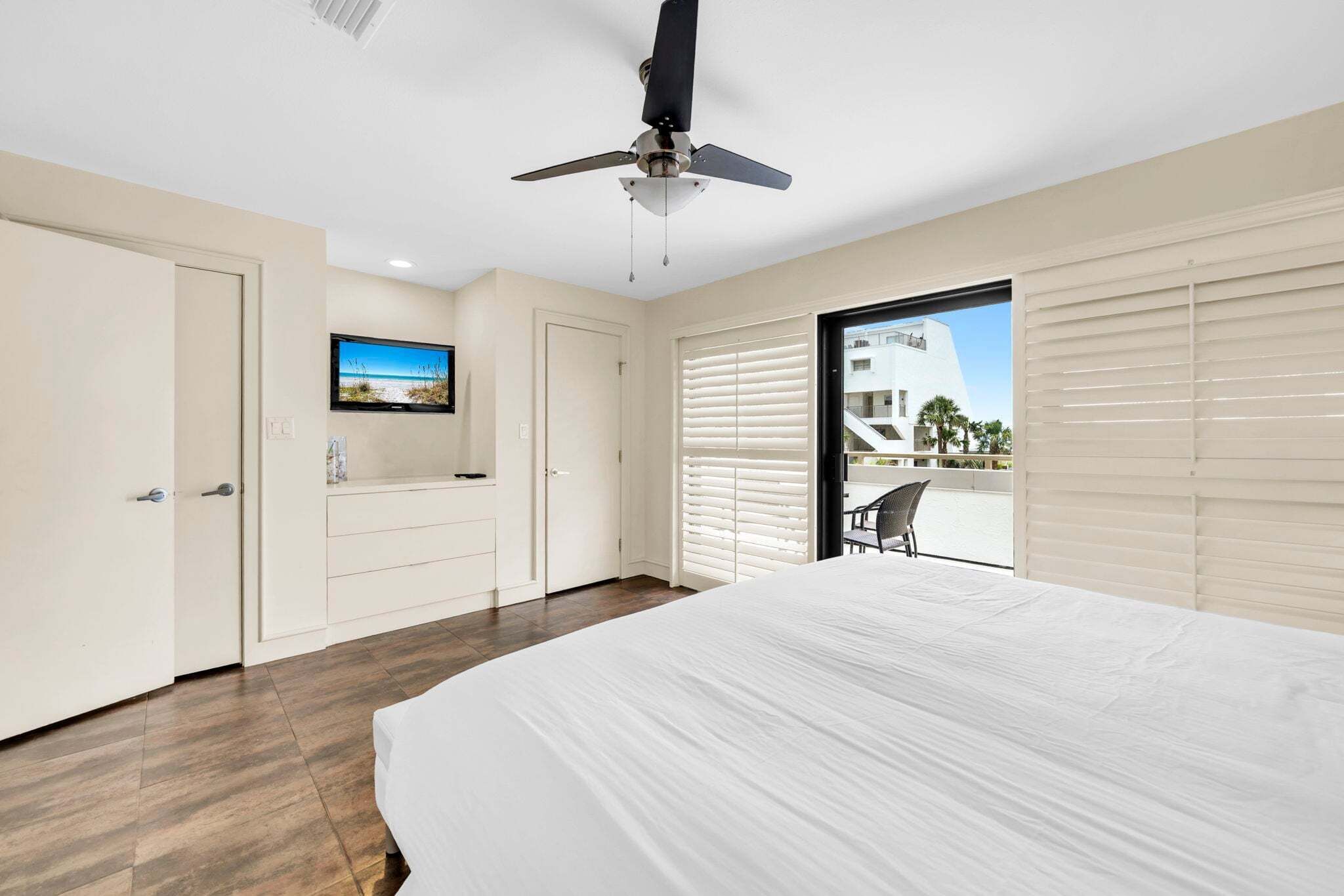 Ein Schlafzimmer mit Bett, Deckenventilator, Fernseher, einer Schiebetür und Blick auf ein Gebäude.
