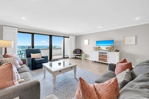 A modern living room with a sofa, coffee table, and a television. There is a balcony with a view of the ocean.