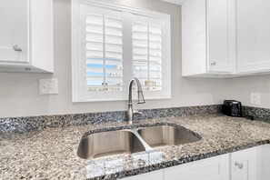 Una cocina con un lavamanos de acero inoxidable doble, encimera de granito, armarios blancos y una ventana con persianas.