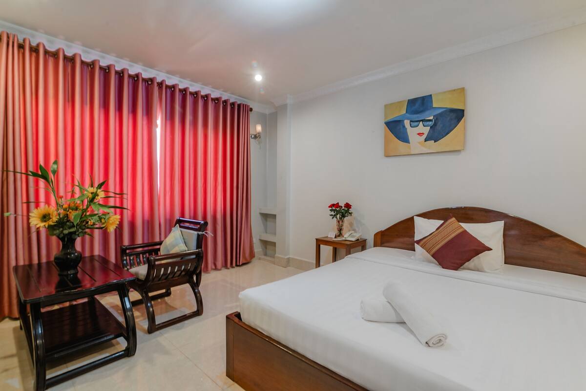Quarto de hotel com cama, escrivaninha com cadeira, um vaso de flores e um quadro na parede.