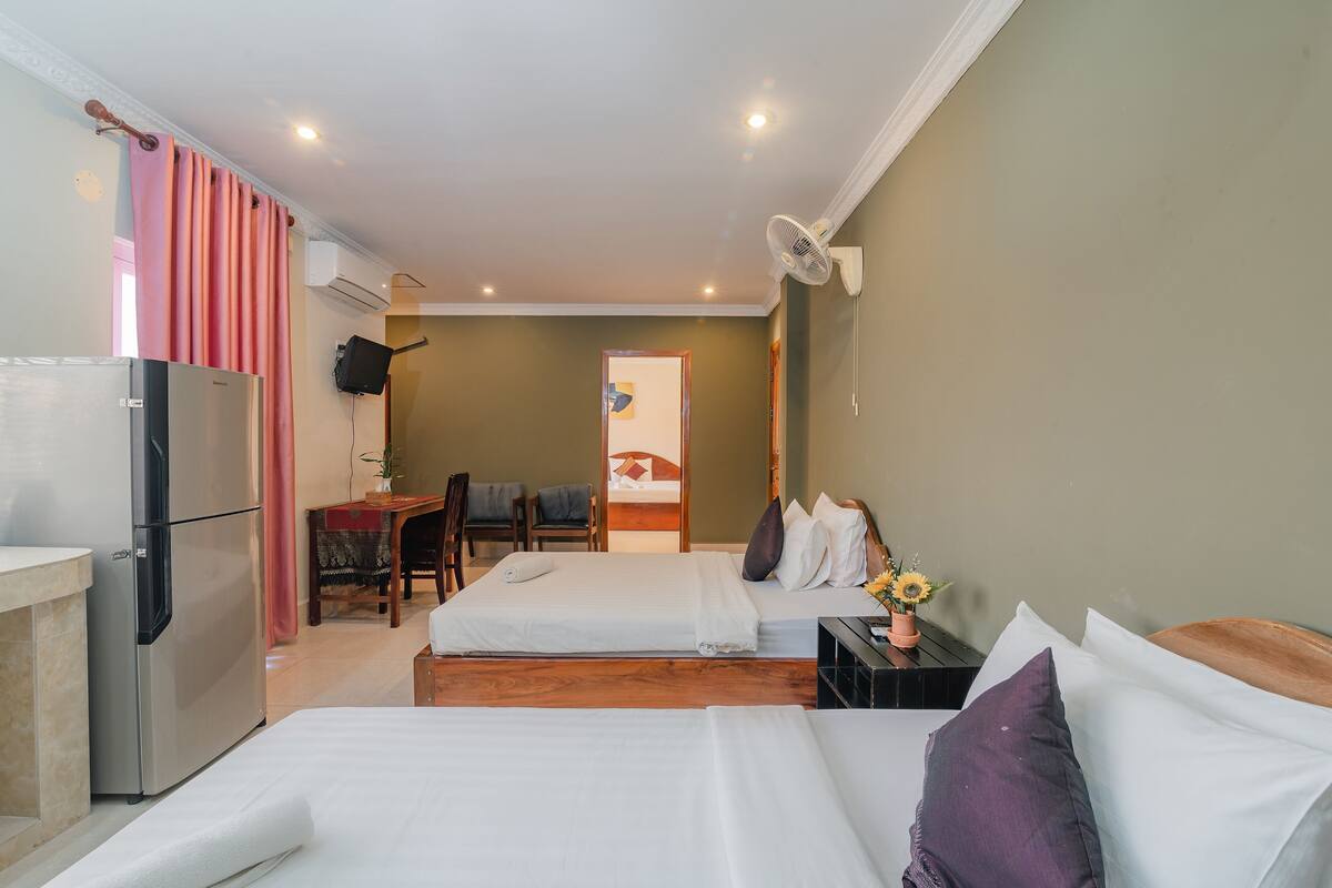 Quarto de hotel com duas camas, uma escrivaninha, uma cadeira, um frigorífico e um ventilador fixado na parede.