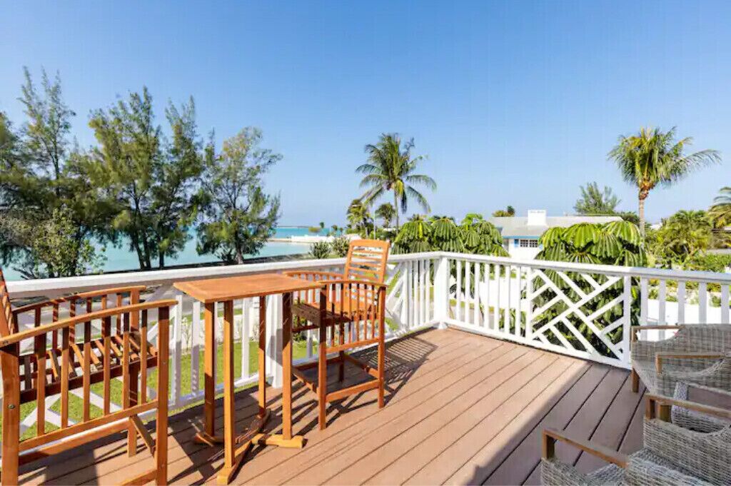 A wooden deck with a table and chairs, overlooking a beach with palm trees and a clear sky.