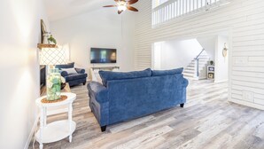 Un salon spacieux avec un canapé bleu, une petite table d’appoint blanche, un ventilateur de plafond et un téléviseur.