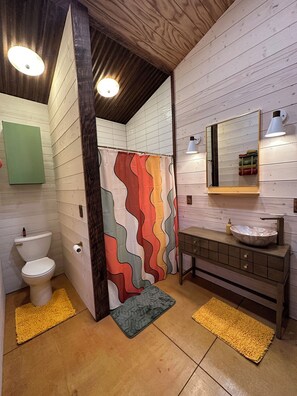 Un bagno con soffitto in legno, un water bianco, una tenda da doccia colorata, un lavabo con bacinella e tappetini gialli.