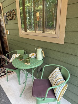 Un tavolo in metallo verde con un bollitore, tazze e un cesto su un portico con sedie e cuscini verdi.