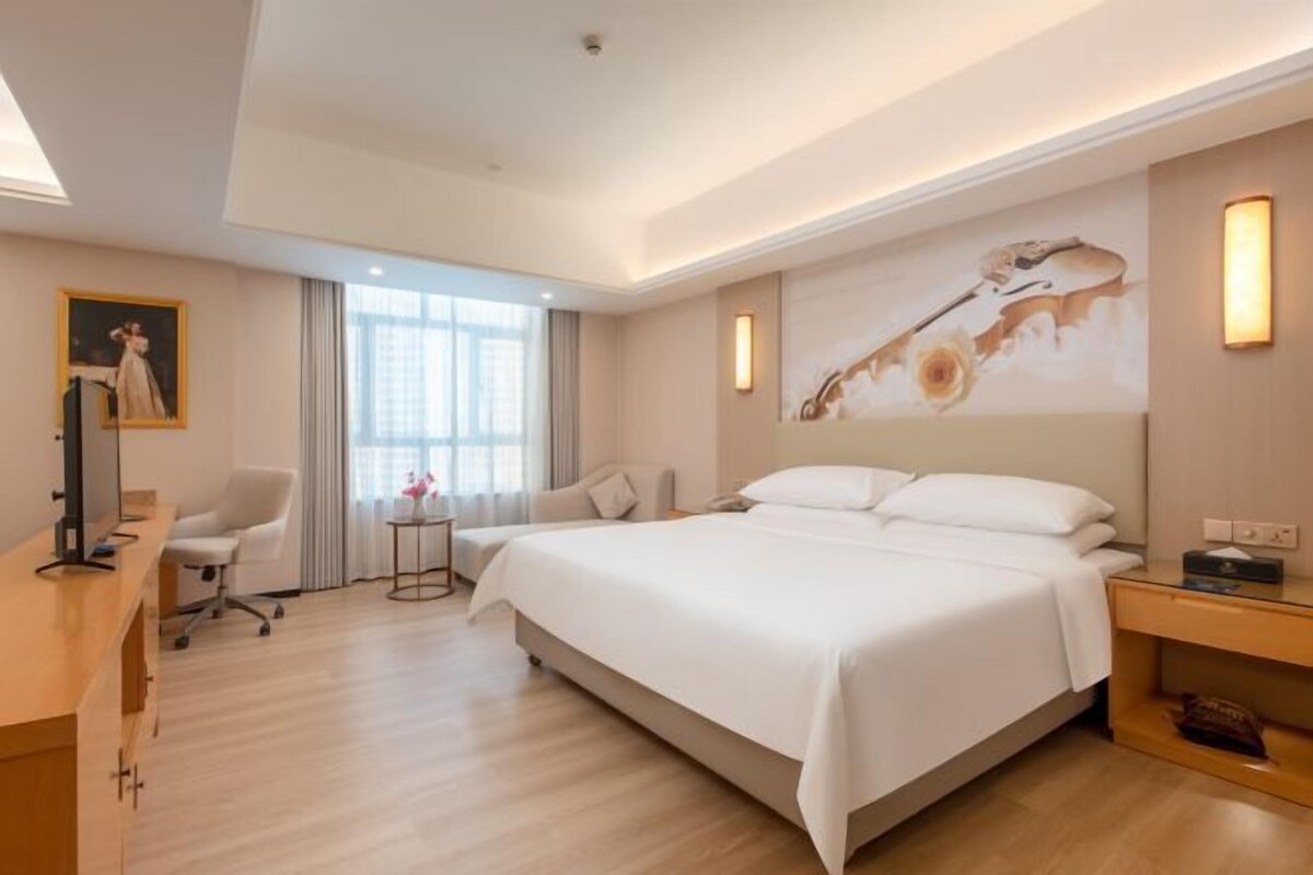A hotel room with a large bed, a desk with a chair, a small table with a lamp, and a painting on the wall.
