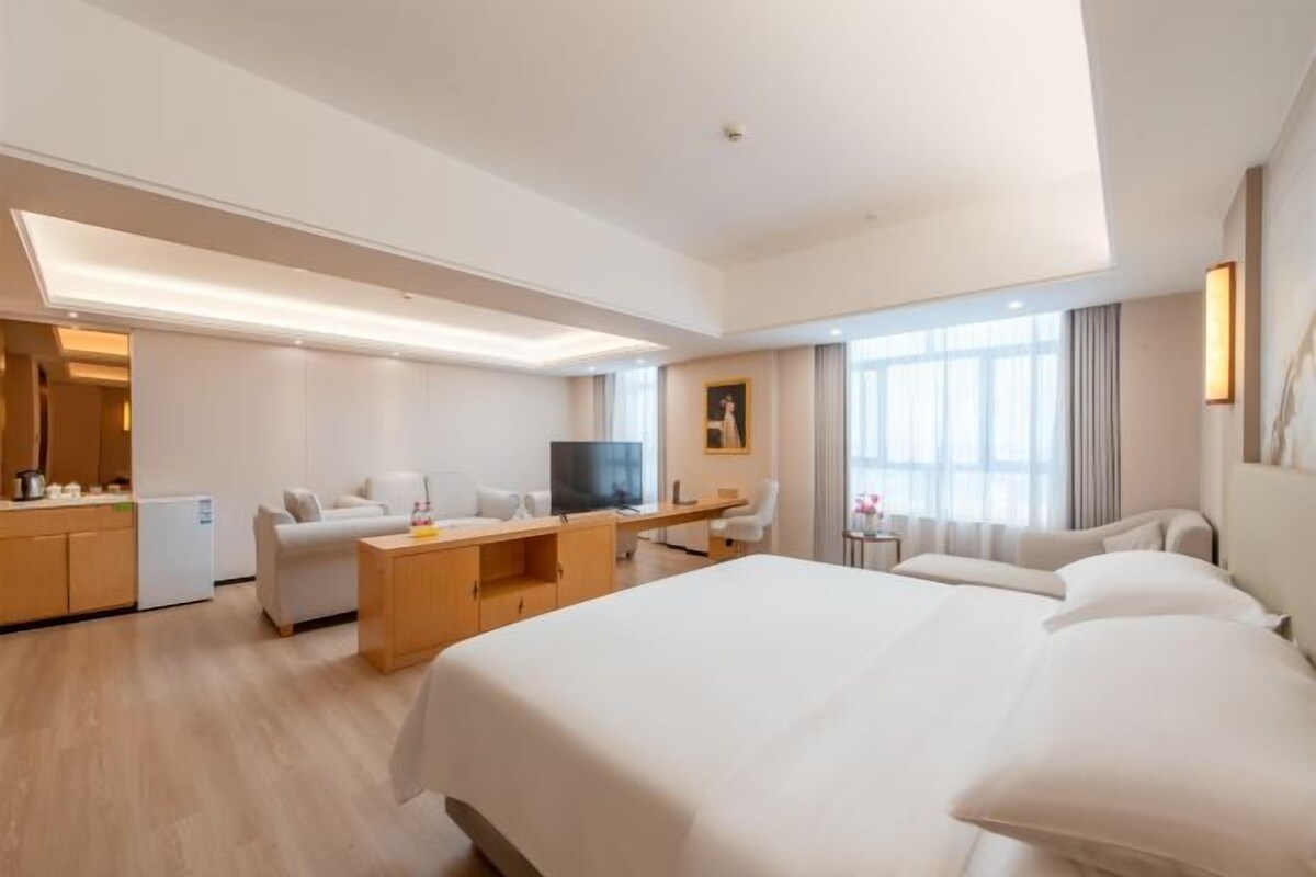A hotel room with a large bed, a desk, a chair, a TV, and a kitchenette.