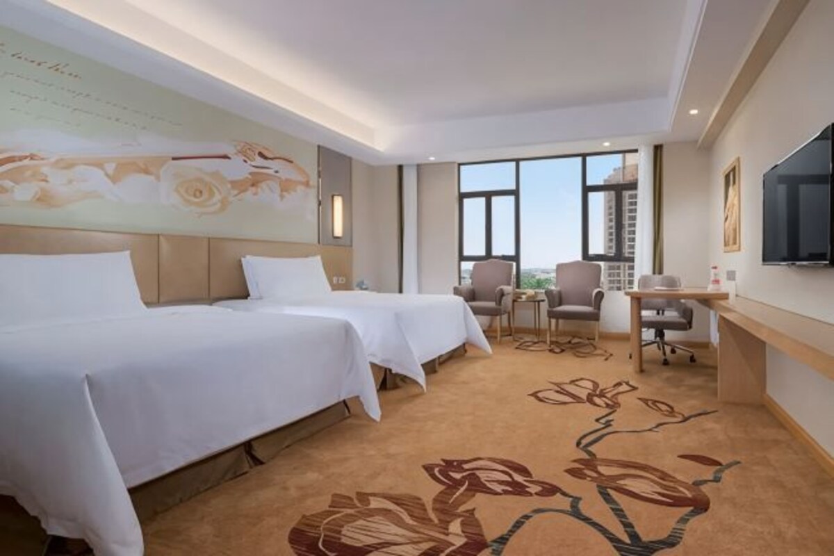 A hotel room with two beds, a desk, a chair, a TV, and a large window.