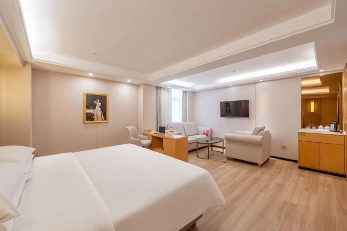 A hotel room with a large bed, a desk with a chair, a sofa, a TV, and a bathroom area.