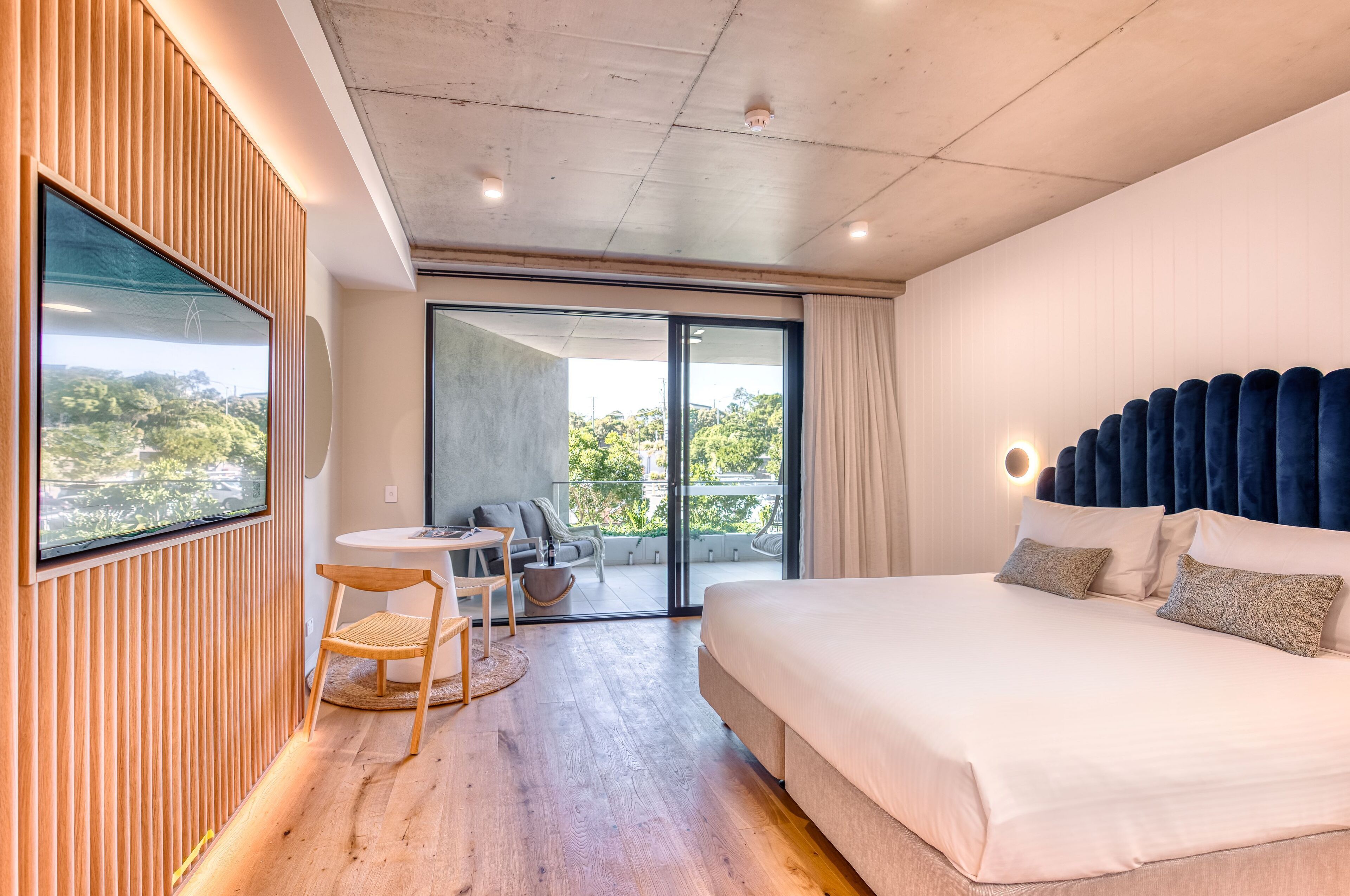 A modern hotel room with a large bed, a balcony with a view, a seating area, and a TV on the wall.