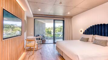 A modern hotel room with a large bed, a balcony with a view, a seating area, and a TV on the wall.