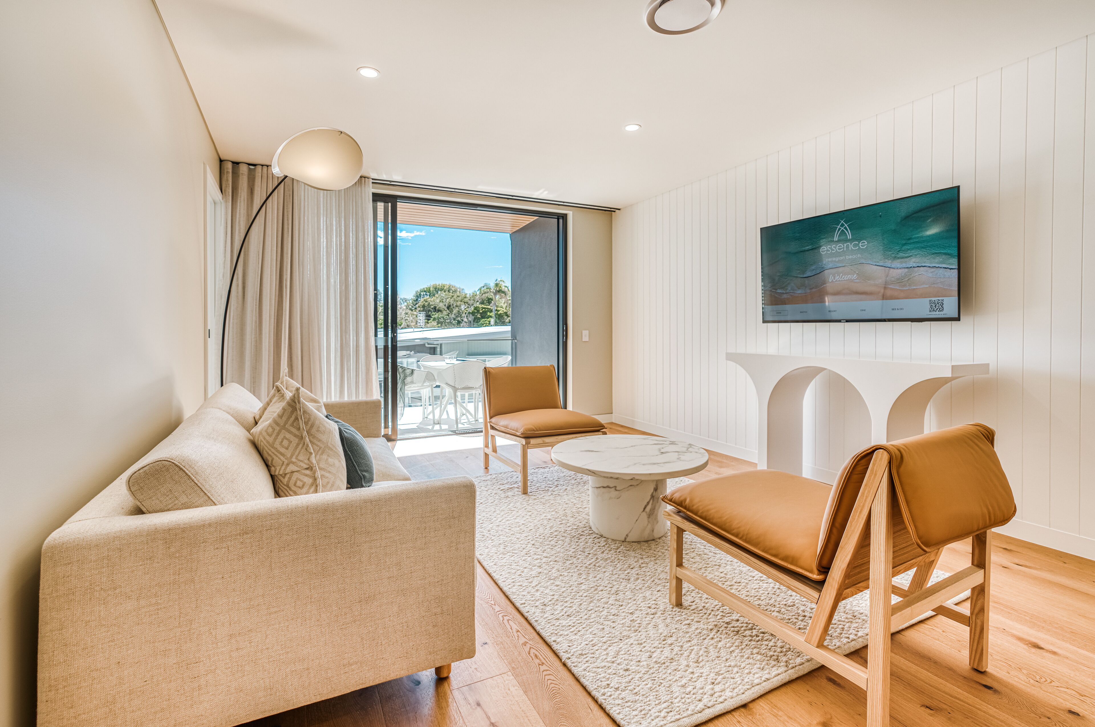 Ein modernes Wohnzimmer mit einer Couch, einem Couchtisch und zwei Sesseln. Es gibt ein großes Fenster mit Vorhängen, einen Flachbildfernseher an der Wand und einen Blick ins Freie.