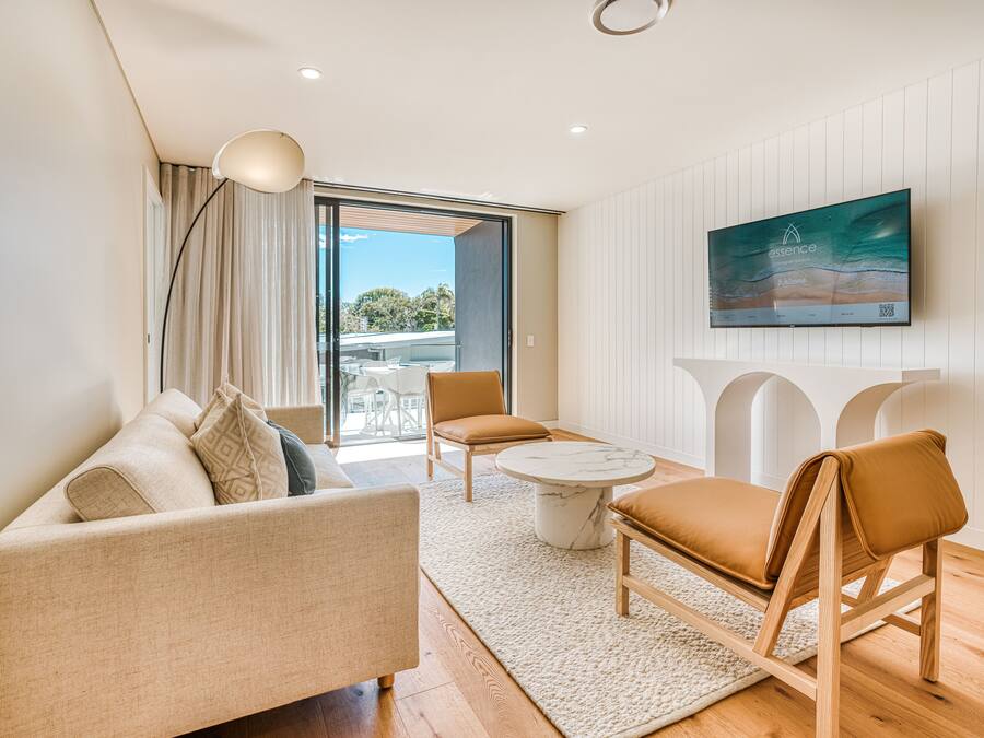Ein modernes Wohnzimmer mit einer Couch, einem Couchtisch und zwei Sesseln. Es gibt ein großes Fenster mit Vorhängen, einen Flachbildfernseher an der Wand und einen Blick ins Freie.