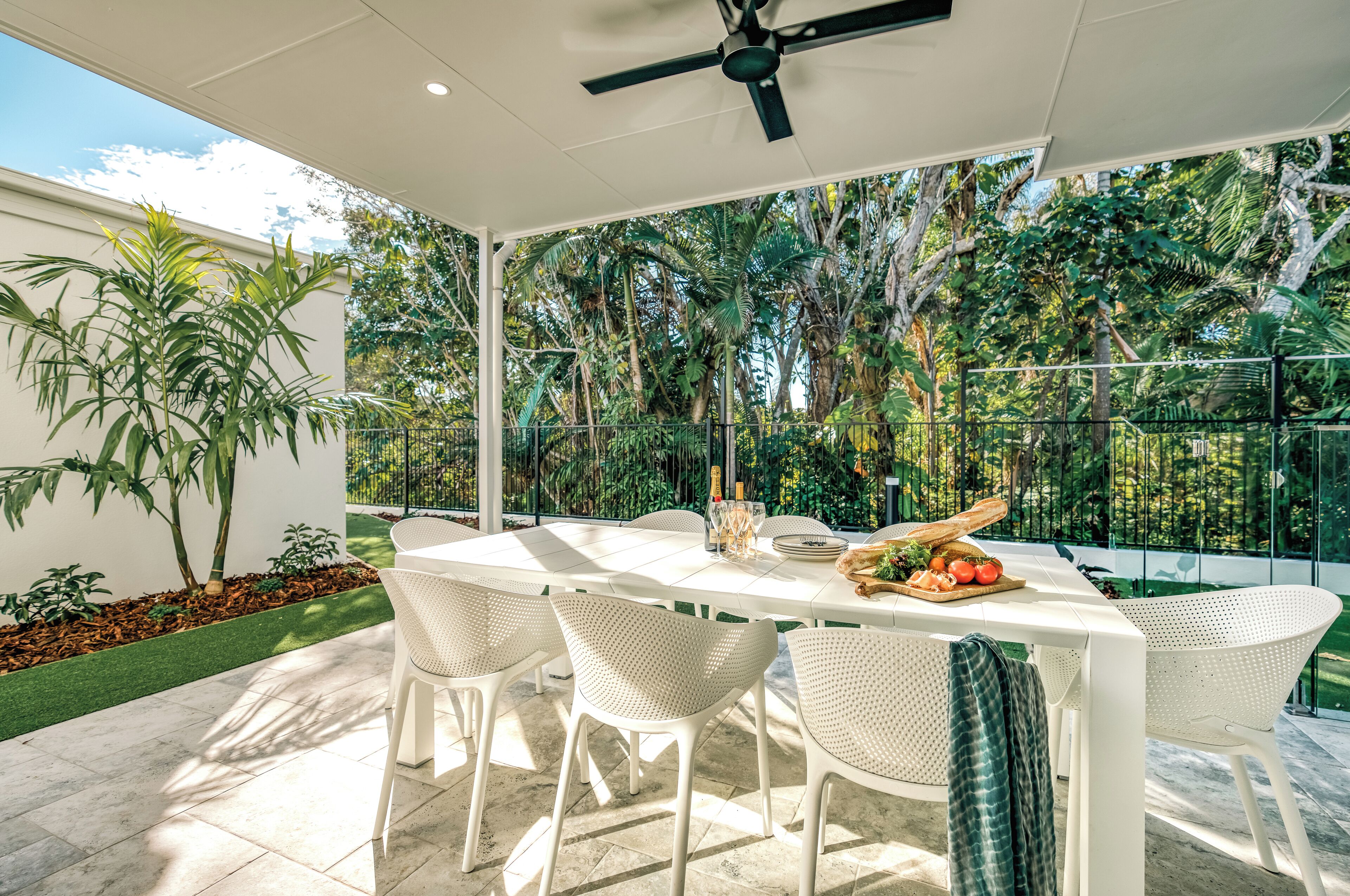 A patio with a white table and chairs, surrounded by lush greenery and a glass fence.