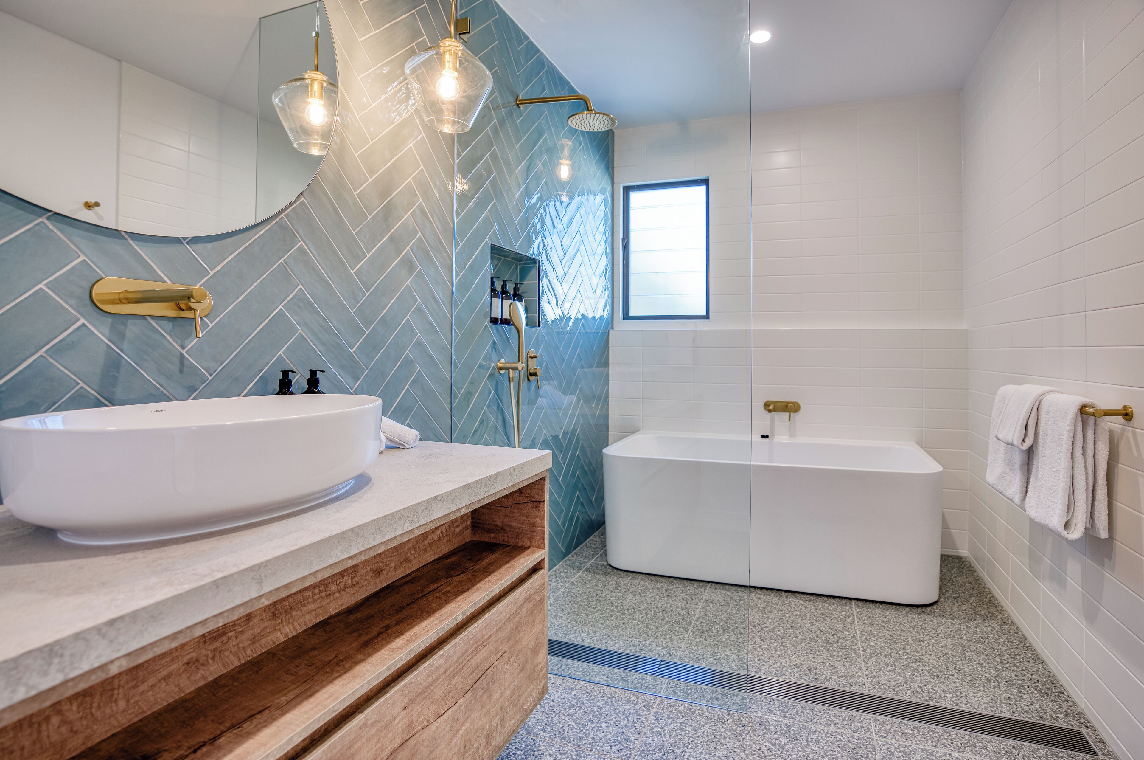 A modern bathroom with a large white bathtub, a wooden vanity with a white sink, and a mirror with a herringbone tile backsplash.