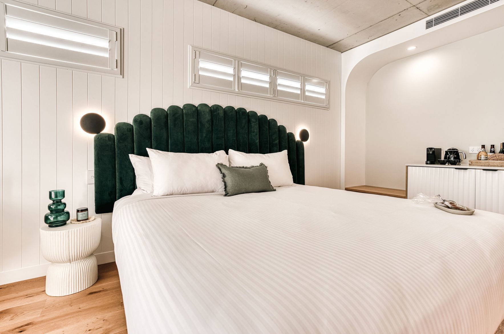 Un dormitorio moderno con una cama grande, un cabecero tapizado en verde, mesitas de noche y una pequeña zona de kitchenette.