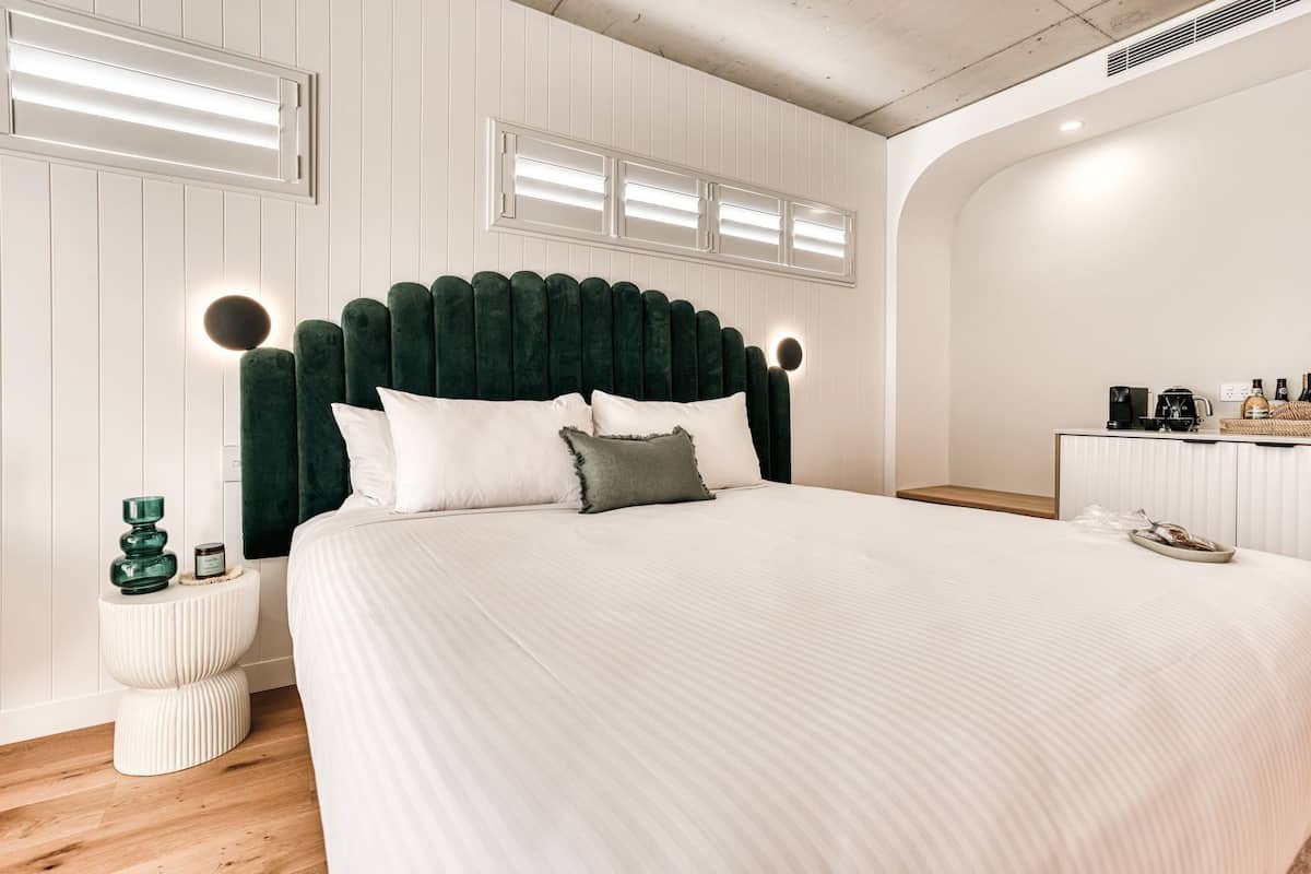 A modern bedroom with a large bed, a green upholstered headboard, bedside tables, and a small kitchenette area.
