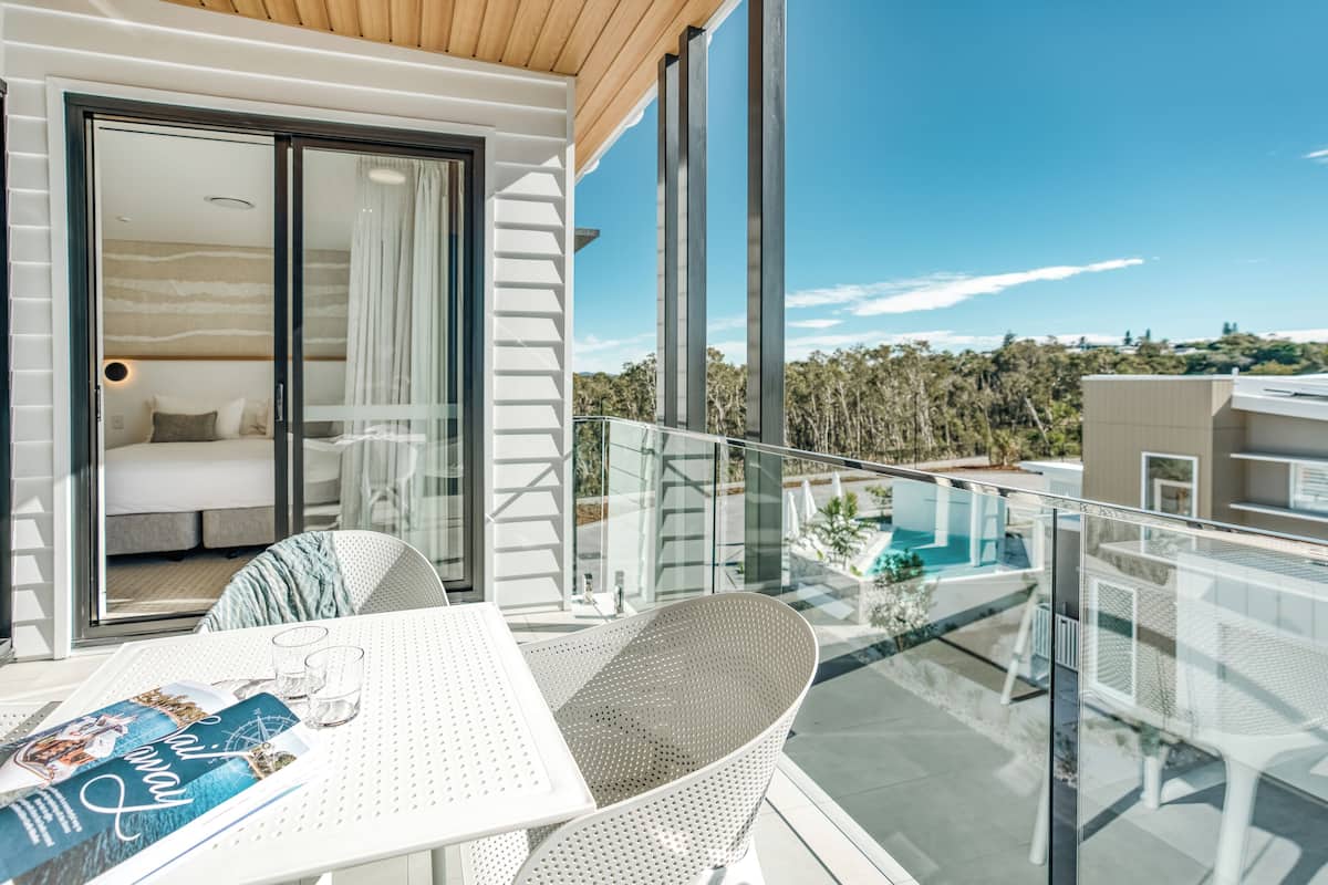 A balcony with a glass railing, a white table, and a chair. There is a bed with a view through the glass door, and a pool in the background.