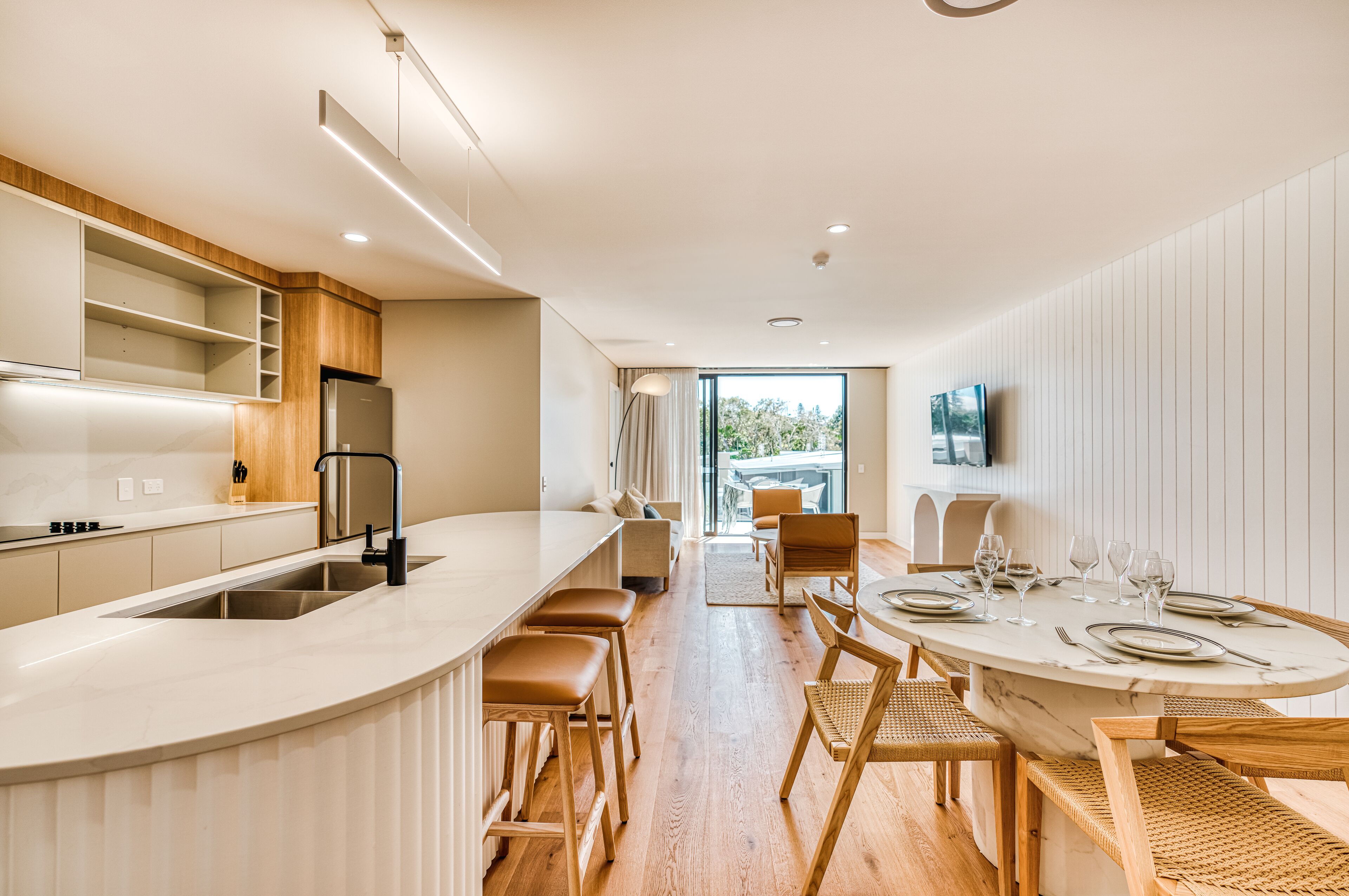 Une cuisine moderne dotée d’un îlot central aux lignes courbes, de meubles en bois et d’un coin repas avec une table en marbre et des chaises en osier.