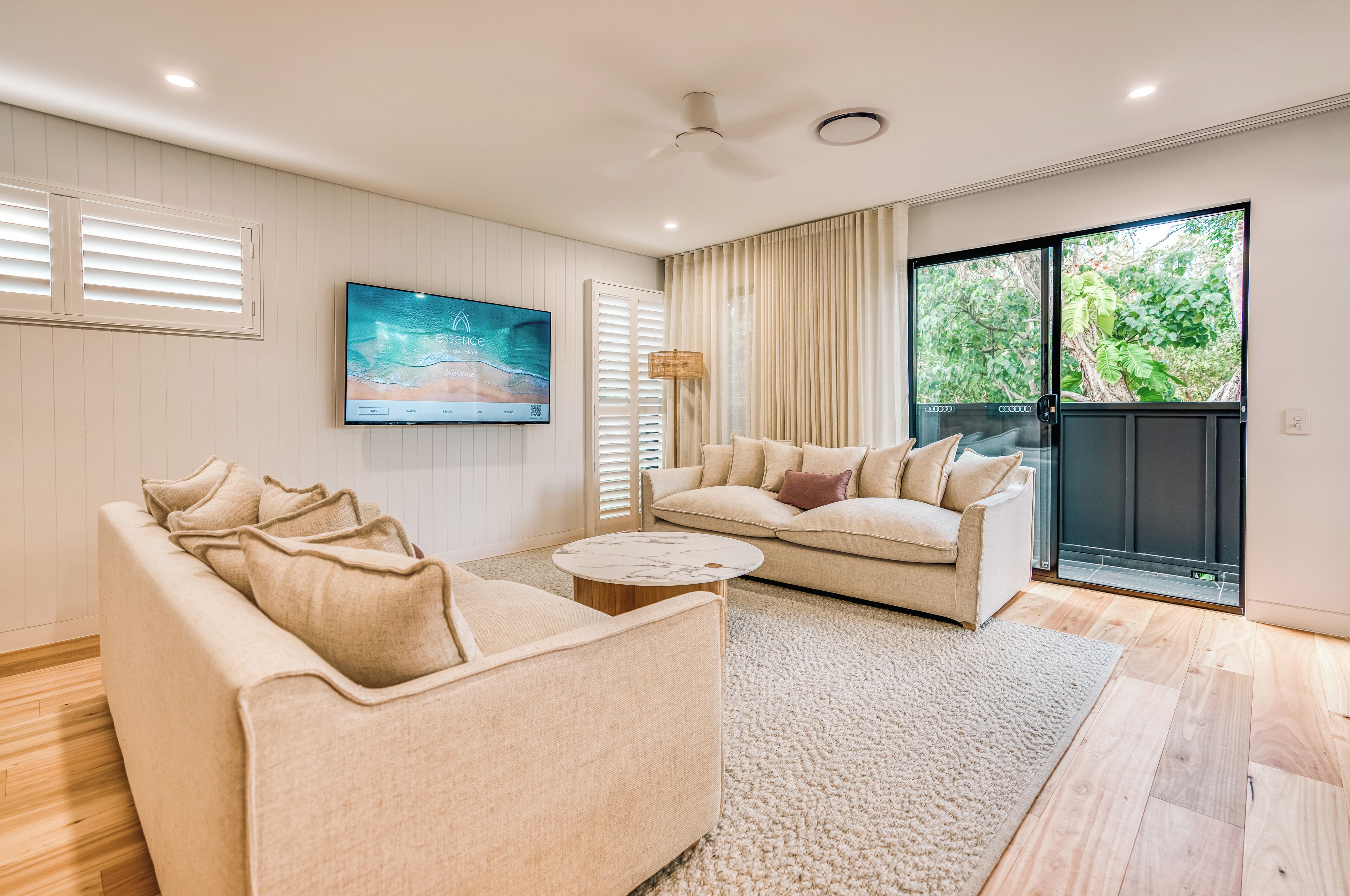A modern living room with a beige sofa, a round coffee table, and a large flat-screen TV mounted on the wall. There is a sliding glass door leading to a balcony with a view of greenery.