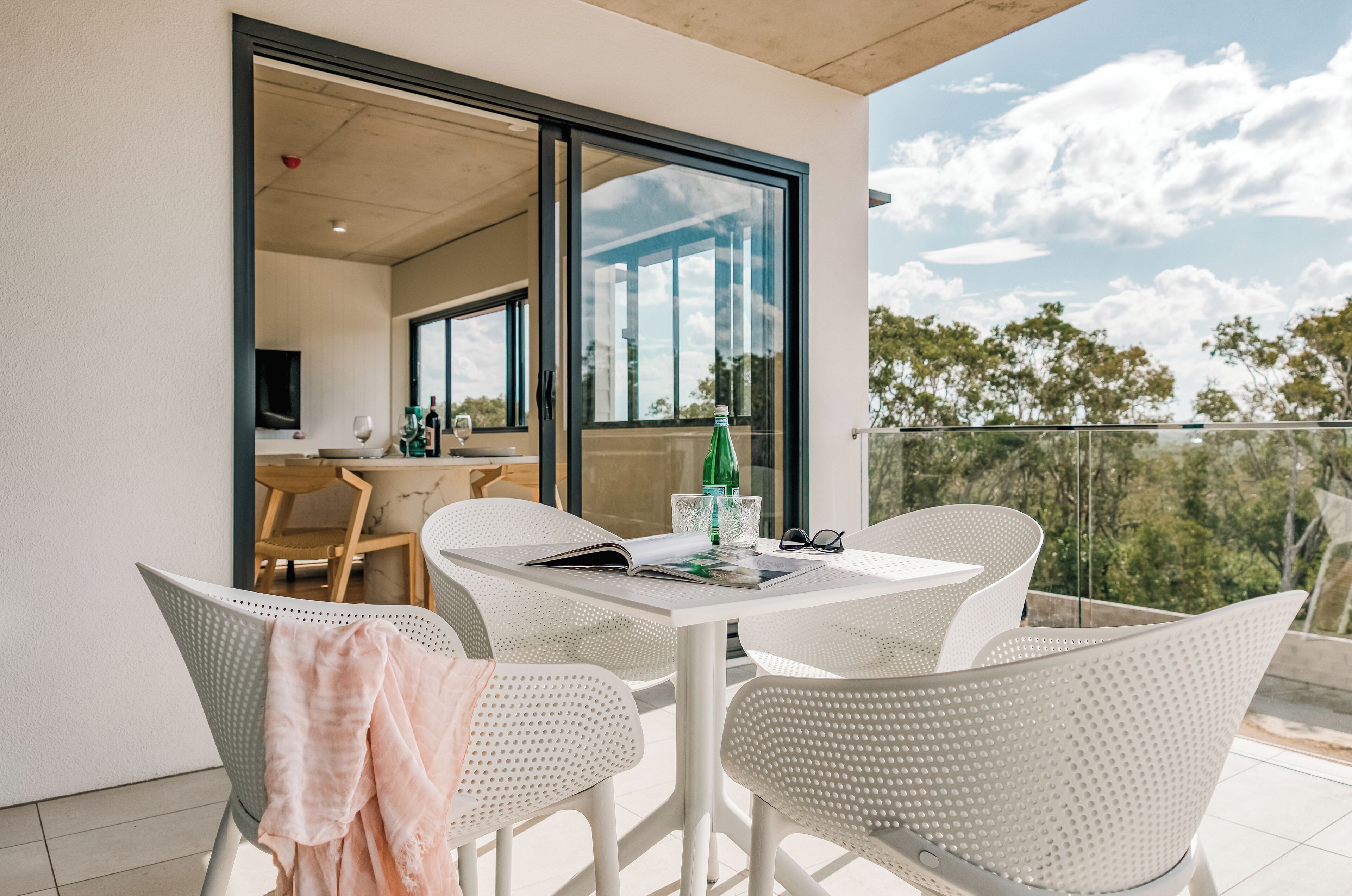 A balcony with a dining table and chairs, a glass door leading to an interior space, and a view of trees and sky.