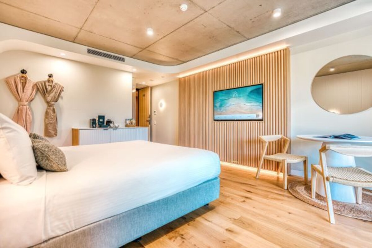 Un dormitorio moderno con una cama grande, un escritorio con una silla, un televisor montado en un panel de madera y un espejo redondo en la pared.