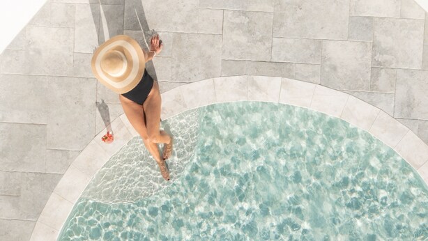 Una persona con traje de baño negro y sombrero de paja, de pie al borde de una piscina con agua cristalina.